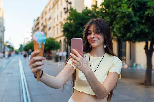Amable hembra con delicioso helado en cono de gofre tomando fotos en el teléfono celular en el pavimento urbano - foto de stock