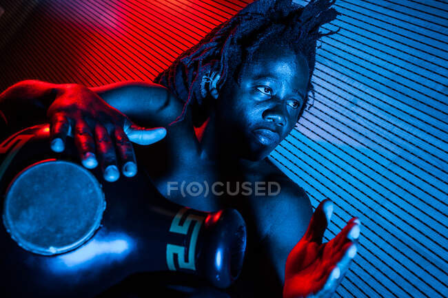 Soñador músico negro masculino con torso desnudo tocando tambor africano en estudio con luces de neón rojas y azules - foto de stock