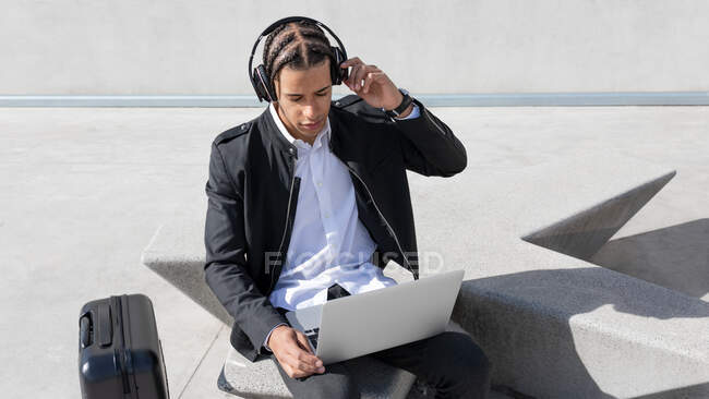 Junge, fokussierte afroamerikanische männliche Manager mit Zöpfen im formellen Anzug und drahtlosen Kopfhörern bei der Arbeit am Laptop auf der städtischen Straße — Stockfoto