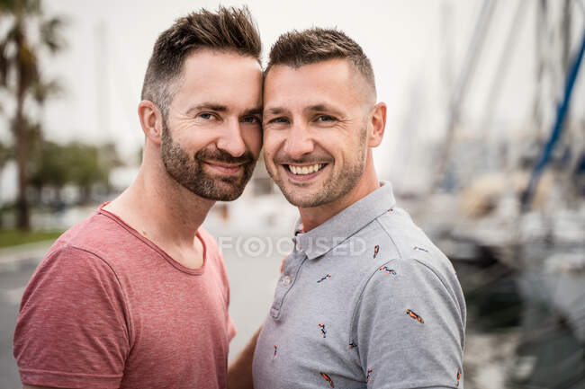 Zufriedenes Paar homosexueller Männer in T-Shirts, die sich umarmen, während sie in die Kamera in der Stadt schauen — Stockfoto