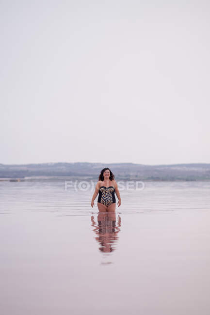 Delighted curvy female in bikini standing in water of pink pond in summer and looking at camera — Stock Photo