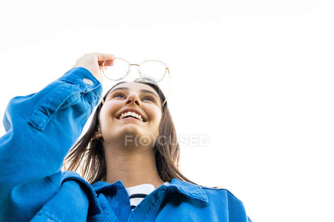 Niedriger Winkel der entzückten Frau mit Brille und blauer Jeansjacke, die auf weißem Hintergrund steht und nach vorne blickt — Stockfoto