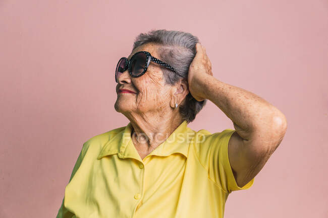 Glückliche moderne ältere Frau mit grauen Haaren und trendiger Sonnenbrille, die den Kopf auf rosa Hintergrund im Studio berührt und wegschaut — Stockfoto