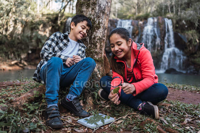 Ethnische Mädchen mit Bruder studiert Pflanzenblätter mit Lupe, während sie gegen Fluss und Kaskade sitzt — Stockfoto