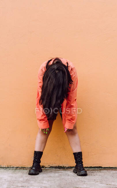 Corps complet de jeune femme anonyme couvrant le visage avec de longs cheveux bruns se penchant vers l'avant tout en se tenant contre un mur orange — Photo de stock