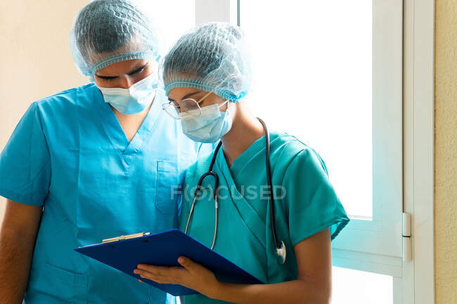 Serious male and female medics in masks reading report on clipboard while standing in corridor in clinic — Stock Photo