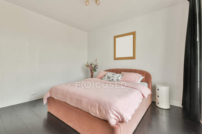 Домашний интерьер просторной спальни с белыми стенами и деревянным полом, обставленной удобной кроватью с розовым покрывалом и подушками и украшенной цветами и макетом картины при дневном освещении — стоковое фото
