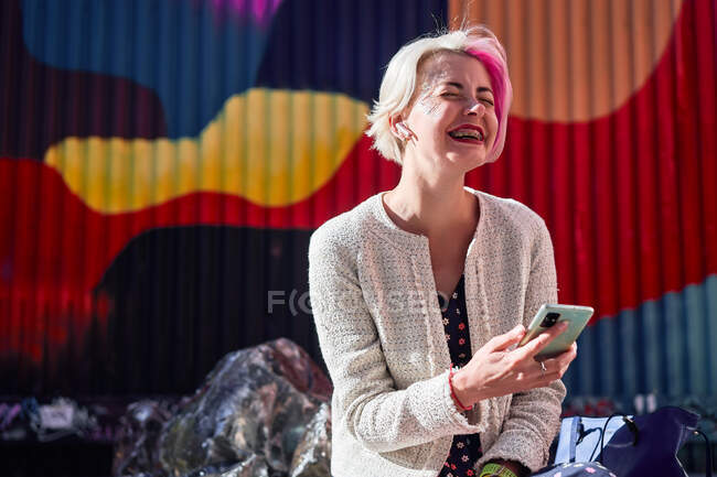 Вид сбоку триумфальной альтернативной женщины с окрашенными волосами, сидящей на скамейке в городском районе и просматривающей социальные сети на мобильном телефоне — стоковое фото