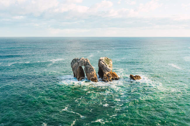 Desde arriba increíble paisaje marino con formaciones rocosas rodeadas de aguas turquesas ondulantes bajo el cielo azul nublado cerca de Liencres en Cantabria España - foto de stock