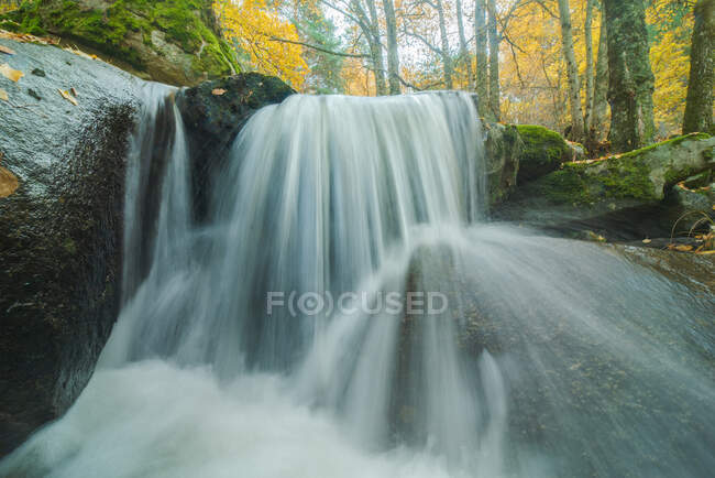 Pittoresca veduta della cascata con fluido d'acqua schiumoso tra massi con muschio e alberi dorati in autunno — Foto stock