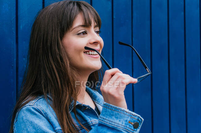 Delighted female with glasses and in denim jacket looking away on background of blue wall in city — Stock Photo