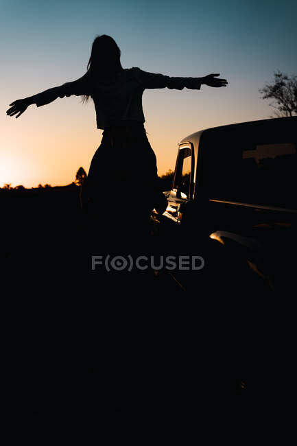 Вид ззаду силует невпізнаваної жінки з простягнутими руками, що сидять на плечах людини, що стоїть біля машини в природі на тлі сонячного неба — стокове фото