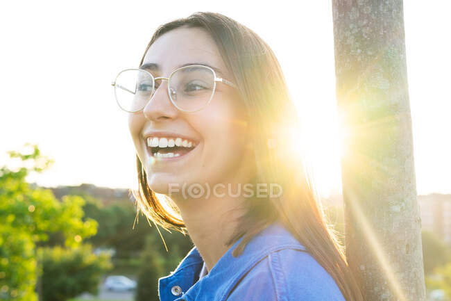 Side view of cheerful female in stylish outfit leaning on tree trunk while standing looking away on green lawn against city in back lit — Stock Photo