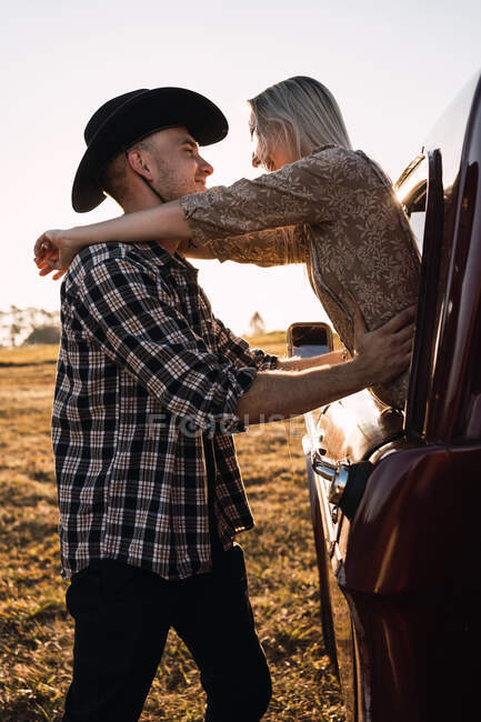 Vista lateral da mulher encantada saindo da janela do carro vintage e abraçando o namorado na camisa xadrez e chapéu de cowboy ao pôr do sol no campo — Fotografia de Stock
