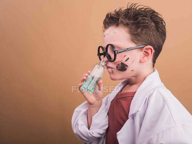 Chemist child in plastic glasses with paint spots on face smelling liquid from bottle on beige background — Stock Photo