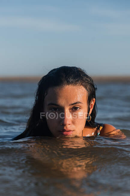 Serena hembra con el pelo mojado nadando en el mar tranquilo en la noche de verano y mirando a la cámara - foto de stock