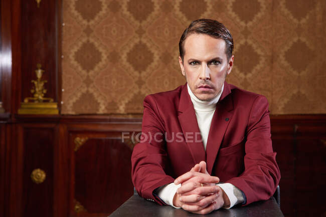 Элегантный стильный театральный мужчина в костюме в студии в стиле ретро смотрит в камеру, сидя на столе — стоковое фото
