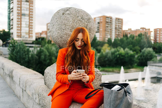 Stylish female with red hair and in vibrant orange suit sitting on stone border in city and messaging on cellphone — Stock Photo