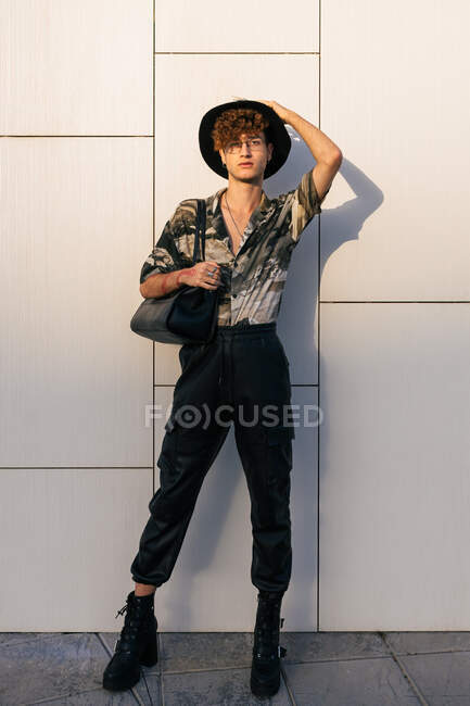 Молодой тщеславный мужчина в стильной одежде с дамской сумочкой, стоящий на черепичной стене, глядя в камеру — стоковое фото