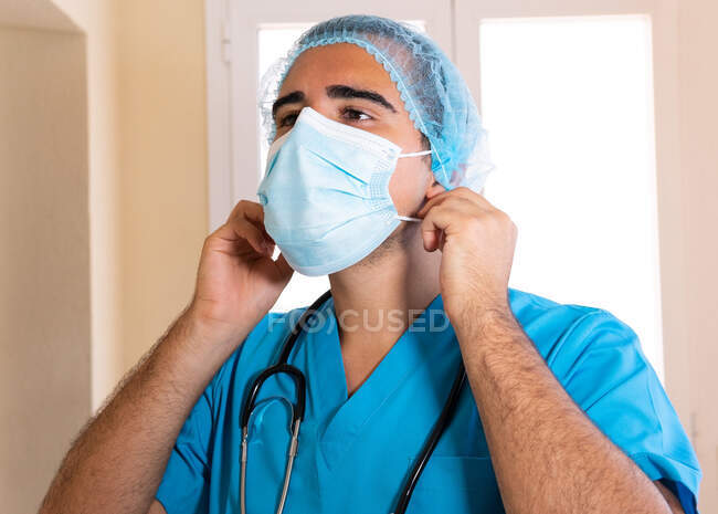 Vista lateral do médico masculino em uniforme colocando máscara descartável durante o trabalho na clínica — Fotografia de Stock