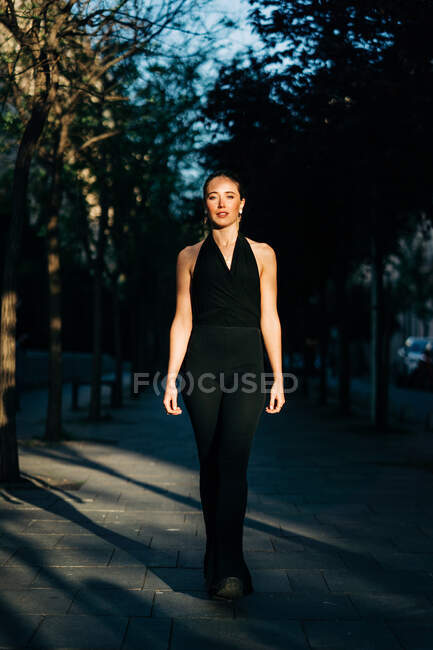 Вид спереду щасливої жінки в модному одязі, що стоїть в місті в сонячний день і дивиться на камеру — стокове фото