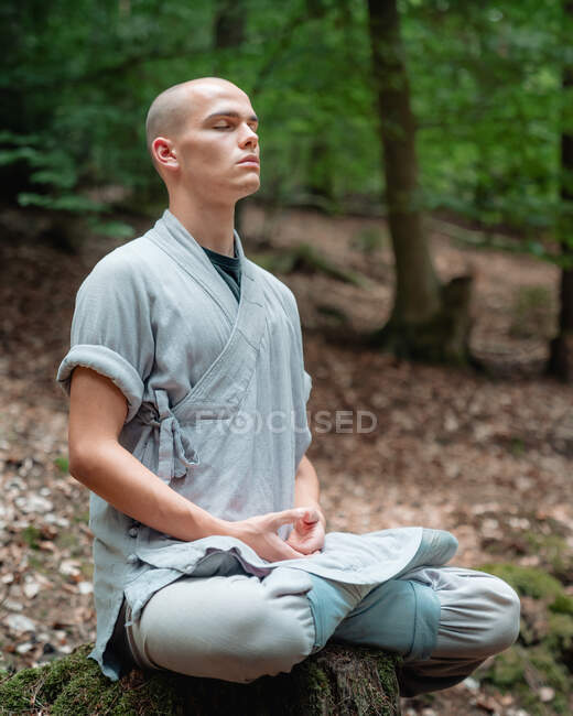 Bald man in traditional clothes sitting on rock in Lotus pose and meditating during kung fu training in forest — Stock Photo