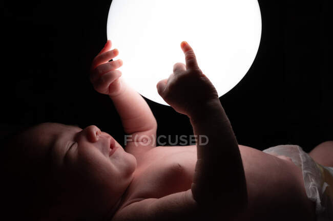 Side view of tender infant sleeping on bed and touching glowing night light lamp in dark room — Stock Photo