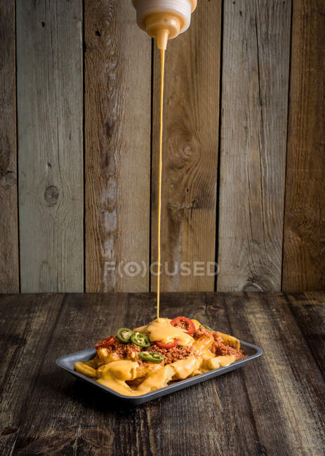 Sauce au fromage à verser sur une assiette avec des frites appétissantes et des poivrons épicés placés sur une table en bois au restaurant — Photo de stock