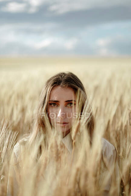 Молодая женщина с волнистыми волосами смотрит на камеру в сельской местности под облачным небом на размытом фоне — стоковое фото