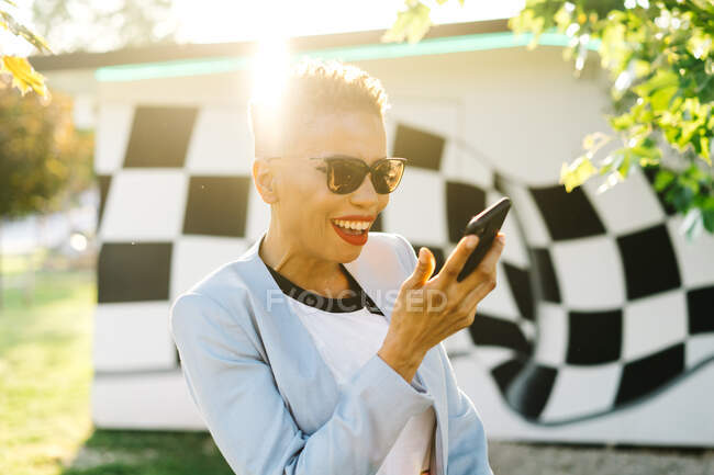 Content mature African American female in modern sunglasses having video chat on cellphone in park in back lit — Stock Photo
