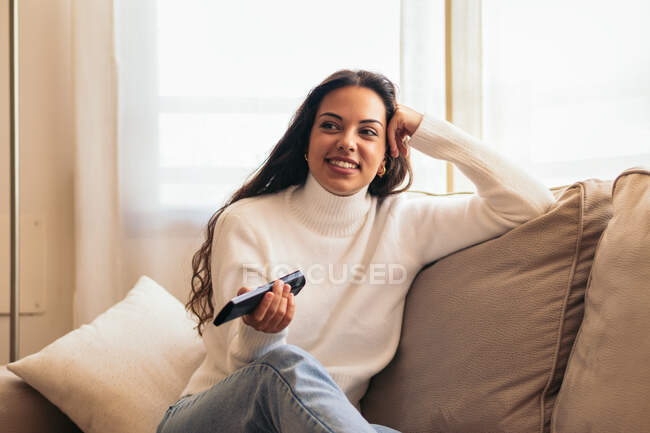 Souriant jeune ethnique femelle changer de canaux sur la télécommande tout en étant assis contre tasse de café et impatient à la maison — Photo de stock