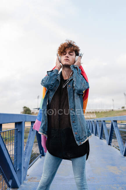 Maschio omosessuale elegante con bandiera LGBT colorata in piedi sul ponte e ascoltare musica in cuffia mentre gli occhi chiusi — Foto stock