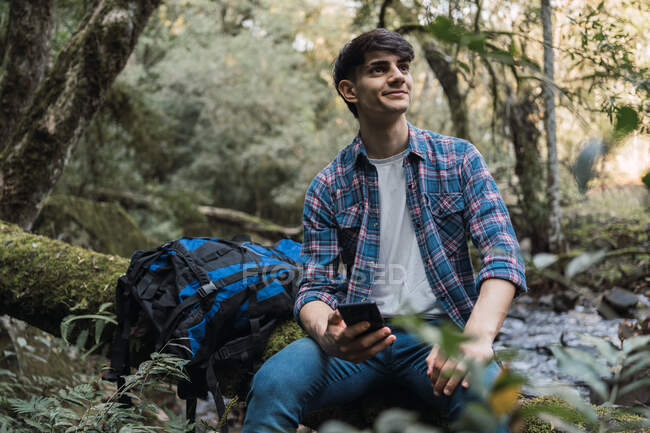 Seitenansicht von zufriedenen männlichen Wanderer mit Rucksack sitzt in der Nähe des Sees im Wald und surft Handy während der Reise — Stockfoto