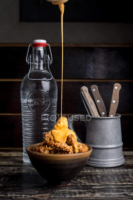 Molho de queijo derramando em delicioso frango crocante colocado na placa perto de garrafa de vidro de água no restaurante escuro — Fotografia de Stock