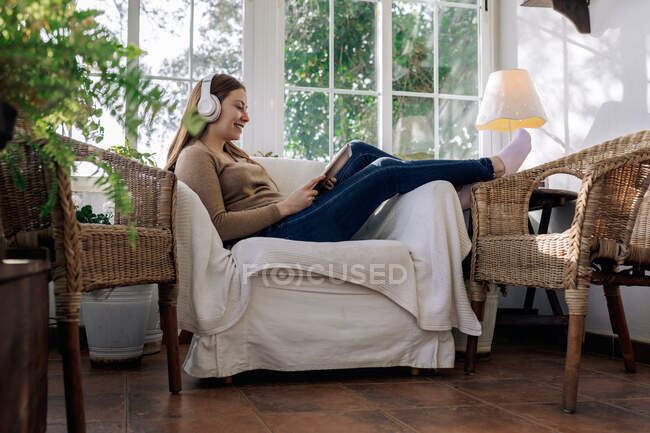 Side view of cheerful young female with tablet listening to music from headphones while sitting in armchair at home — Stock Photo