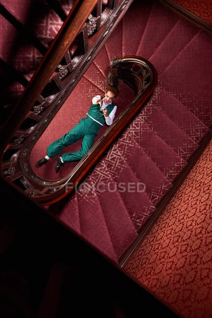 Vista superior de talentoso actor masculino tendido en el suelo cerca de la escalera y fingiendo estar muerto alcanzando mirando a la cámara durante la actuación creativa - foto de stock