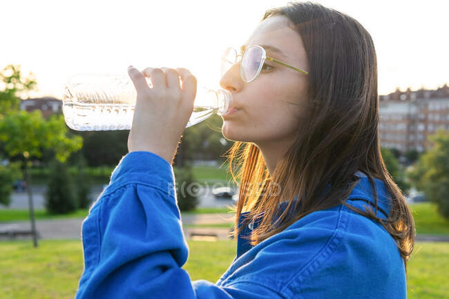 Vista lateral de hembra sedienta con los ojos cerrados bebiendo agua fresca de la botella de plástico en la ciudad en la parte posterior iluminada - foto de stock