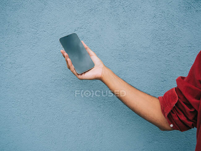 Recorte masculino irreconocible que muestra el teléfono móvil moderno con pantalla negra sobre fondo azul en la ciudad - foto de stock