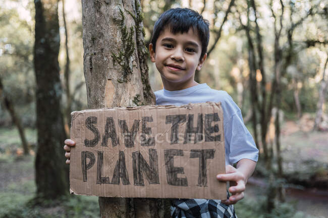 Enfant ethnique souriant montrant le titre Save The Planet sur un carton tout en regardant la caméra dans la forêt — Photo de stock