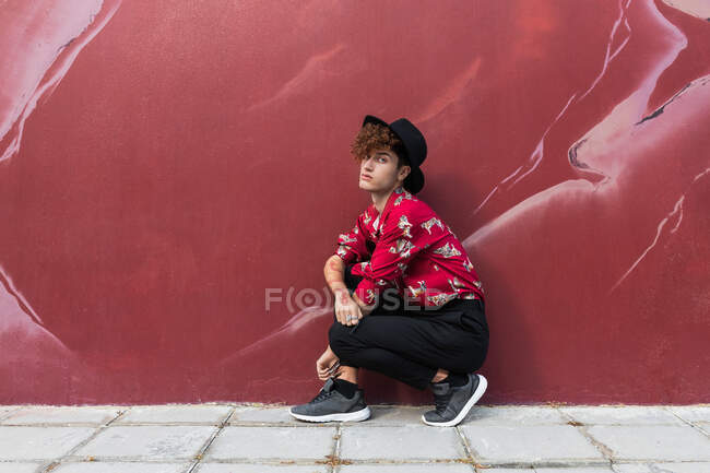 Vue latérale de l'homme homosexuel à la mode avec de longs ongles en chemise ornementale regardant la caméra tout en squattant sur le trottoir contre le mur — Photo de stock