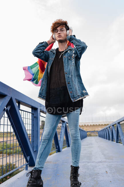 Гомосексуальный стильный мужчина с разноцветным ЛГБТ-флагом, стоящий на мосту и слушающий музыку в наушниках, глядя в камеру — стоковое фото