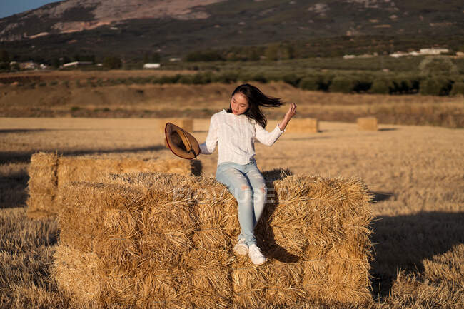 Content ethnic female with straw hat sitting on dry hay stack in field while he touches his hair — Stock Photo