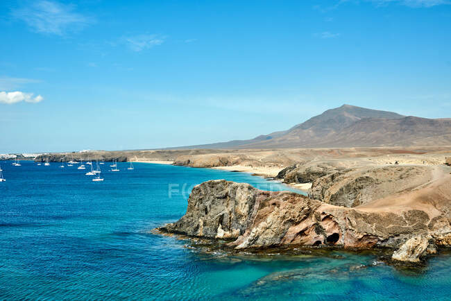 Luftaufnahme der rauen steinigen Klippe in der Nähe des türkisfarbenen Meeres mit Yachten vor blauem Himmel an einem sonnigen Sommertag auf Fuerteventura, Spanien — Stockfoto