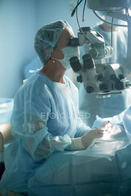Doctora adulta en máscara estéril y gorra médica ornamental mirando a través del microscopio quirúrgico contra compañero de trabajo en el hospital - foto de stock