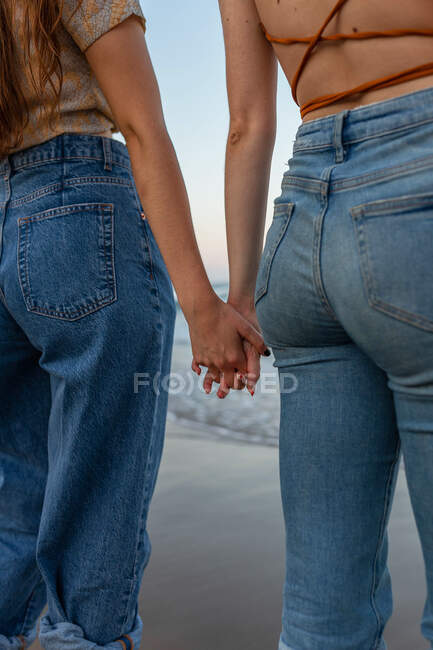Unrecognizable girlfriends in jeans holding hands while standing on wet beach near stormy sea during romantic date — Stock Photo