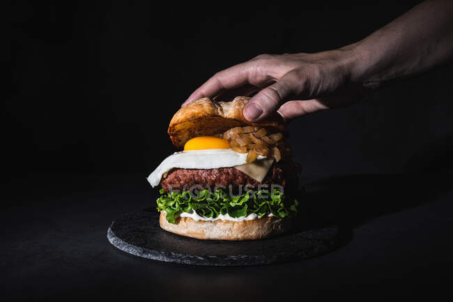 Crop unrecognizable person putting bun on burger with egg and patty served on slate board on black background in studio — Stock Photo