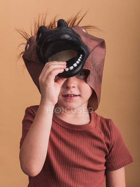 Child in t shirt and horse mask on head looking at camera on beige background — Stock Photo