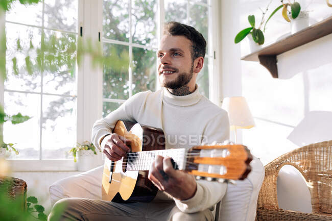 Споглядальний чоловічий музикант з татуюваннями грає на класичній гітарі, сидячи в кріслі і дивлячись у вікно в будинку — стокове фото