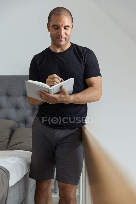 Hombre enfocado de pie cerca de la cama y escribir pensamientos en el diario en la mañana en el dormitorio - foto de stock