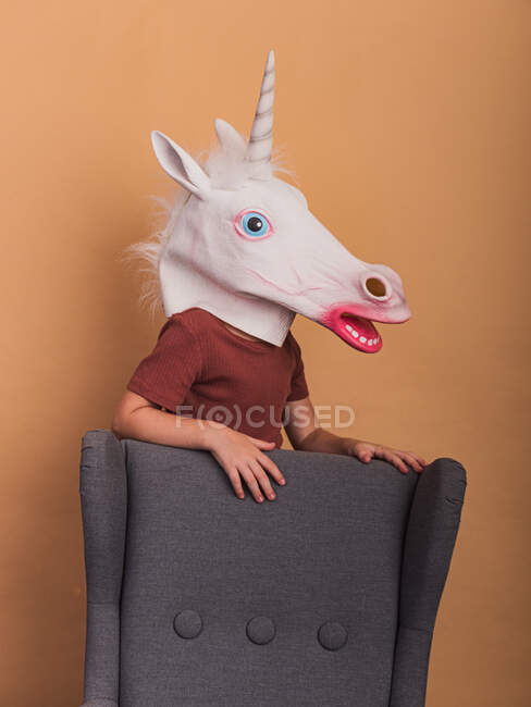 Criança anônima em máscara decorativa unicórnio com boca aberta tocando poltrona no fundo bege — Fotografia de Stock
